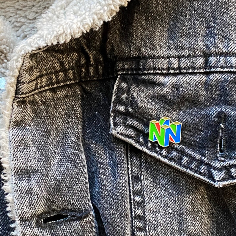 Peut inclure: &Eacute;pinglette color&eacute;e en &eacute;mail repr&eacute;sentant le logo Nintendo 64 vert, rouge et bleu. L'&eacute;pinglette est fix&eacute;e sur une veste en jean noire avec une doublure blanche et duveteuse.