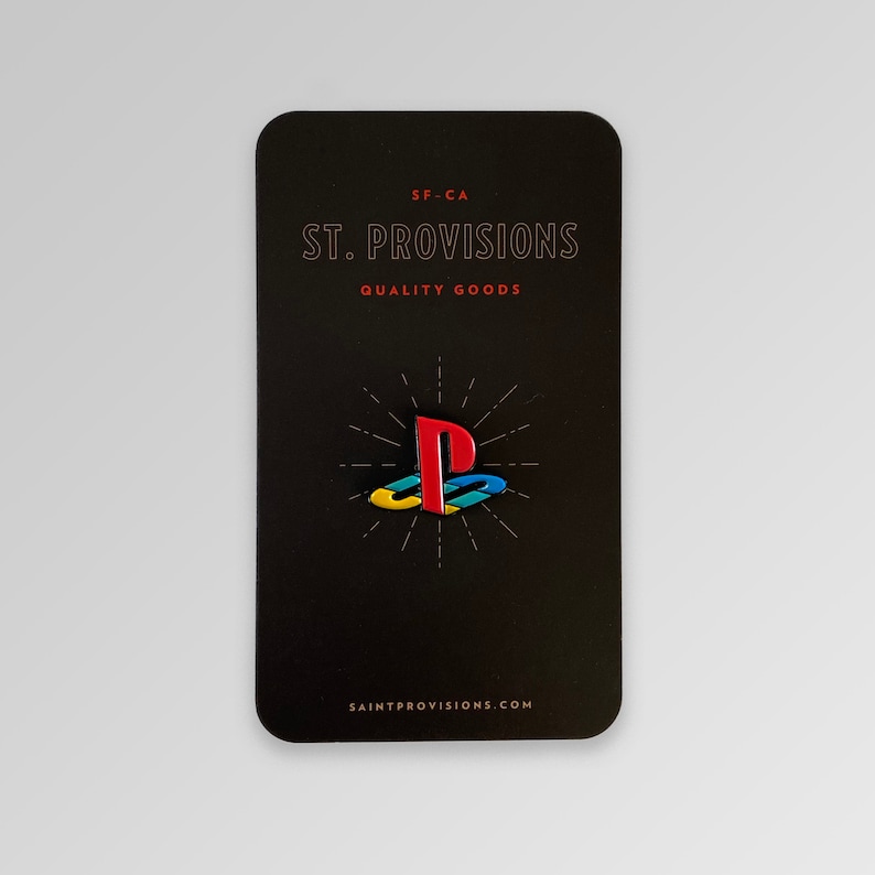 以下が含まれることがあります： PlayStationのロゴが描かれた黒と白のエナメルピン。ピンは、「ST. PROVISIONS QUALITY GOODS」と「SAINTPROVISIONS.COM」のテキストが書かれた黒いカードの上にあります。