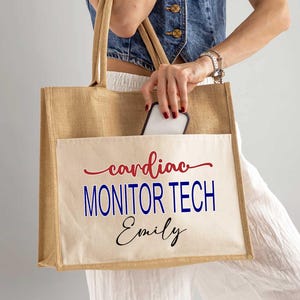 May include: A tan jute tote bag with a front pocket. The pocket features the words "cardiac MONITOR TECH Emily" in red, blue, and black script. The bag has tan handles and is held by a person.