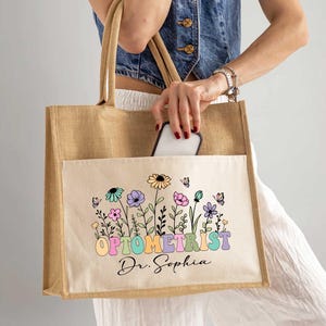 May include: A natural-toned jute tote bag with a front pocket featuring a colorful floral design and the text "OPTOMETRIST Dr. Sophia". The bag has sturdy handles and is being held by a person.