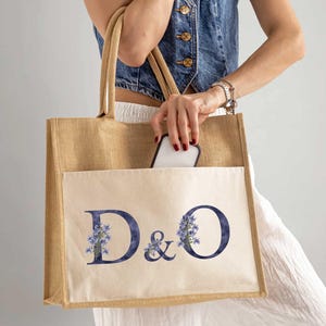 Könnte beinhalten: Beige Tragetasche mit Fronttasche und Henkeln. Die Tasche zeigt die Buchstaben "D&O" in einem dunkelblauen Aquarellstil mit floralen Akzenten. Ein Smartphone ist in der Tasche sichtbar. Die Tasche ist aus einem natürlichen Material.