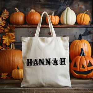 May include: A cream-colored canvas tote bag featuring the name HANNAH printed in black capital letters, with each letter stylized with a spiderweb pattern. The bag has two shoulder straps and is constructed from a plain, woven fabric material.