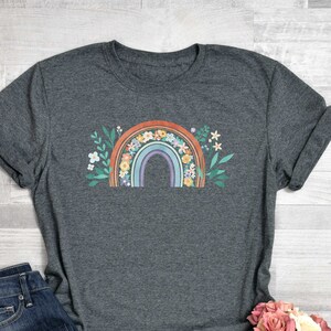 May include: Dark gray heather t-shirt with a rainbow and floral design. The rainbow is multi-colored with orange, red, yellow, green, blue, and purple stripes. The flowers are white, pink, and yellow.