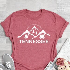May include: Coral pink t-shirt with a white graphic of mountains and trees. The text "Never Stop Exploring" and "Tennessee" are printed on the shirt.