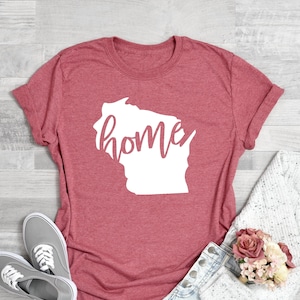 May include: Coral pink t-shirt with a white graphic of the Wisconsin state outline and the word "home" in a cursive font. The shirt is paired with gray sneakers and denim shorts. A white sweater and a bouquet of pink flowers complete the casual ensemble.