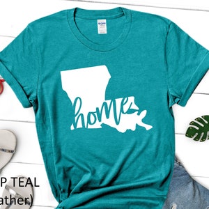 May include: A teal blue t-shirt with a white outline of the state of Louisiana and the word "home" written in a cursive font.