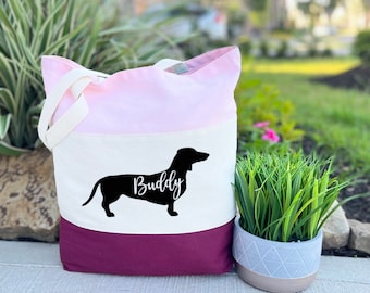 May include: Tri-color tote bag with a pink, white, and burgundy design. The bag features a black silhouette of a dachshund with the name "Buddy" written in white.