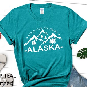 May include: A teal heather t-shirt with a white graphic of mountains and trees. The graphic says "Never Stop Exploring -Alaska-."