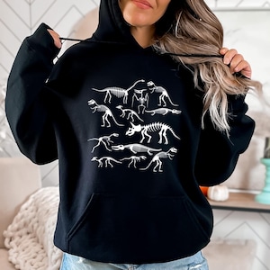 May include: Black hooded sweatshirt with a white graphic print of dinosaur skeletons. The graphic features a variety of dinosaur skeletons in different poses.