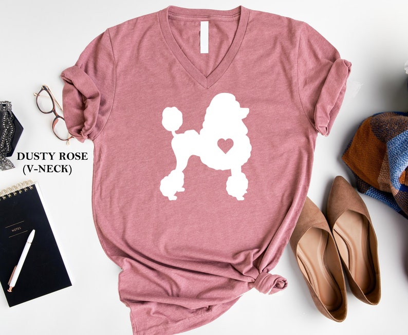 Puede incluir: Camiseta de cuello en V rosa polvoriento con una silueta blanca de un caniche con un coraz&oacute;n en el pecho.