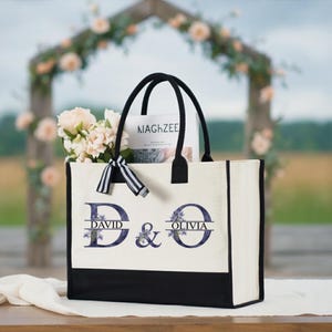 Könnte beinhalten: Eine personalisierte Tragetasche mit schwarzen Griffen und einem schwarzen Boden. Die Tasche trägt die Namen "David & Olivia" in einer stilisierten Schriftart mit floralen Akzenten. Die Tasche ist mit Blumen und einer Zeitschrift gefüllt.