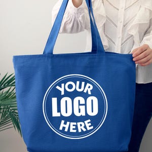 K&ouml;nnte beinhalten: Eine blaue Tragetasche mit langen Henkeln. Die Tasche hat ein wei&szlig;es kreisf&ouml;rmiges Logo mit dem Text "YOUR LOGO HERE". Die Tasche wird von einer Person gehalten, die eine wei&szlig;e Bluse tr&auml;gt.