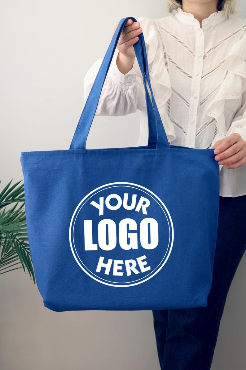 K&ouml;nnte beinhalten: Eine blaue Tragetasche mit langen Henkeln. Die Tasche hat ein wei&szlig;es kreisf&ouml;rmiges Logo mit dem Text "YOUR LOGO HERE". Die Tasche wird von einer Person gehalten, die eine wei&szlig;e Bluse tr&auml;gt.