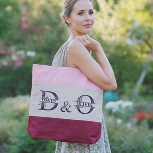 Könnte beinhalten: Eine personalisierte Tragetasche mit rosa Oberteil, beiger Mitte und bordeauxrotem Boden. Die Tasche trägt die Namen "DAVID" und "OLIVIA" in einer dekorativen Schriftart mit floralen Akzenten, getrennt durch ein kaufmännisches Und.