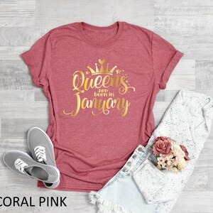 May include: Coral pink t-shirt with gold glitter text that reads "Queens are born in January" and a gold glitter crown above the text.