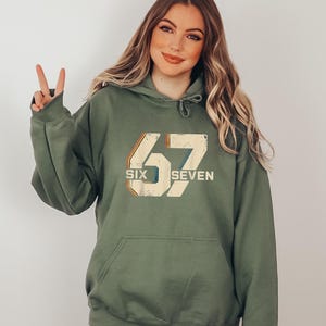 May include: Olive green hoodie with a front pocket and drawstring hood. The front features the number "67" in a retro font, with the words "SIX" and "SEVEN" below. The hoodie is a casual style.