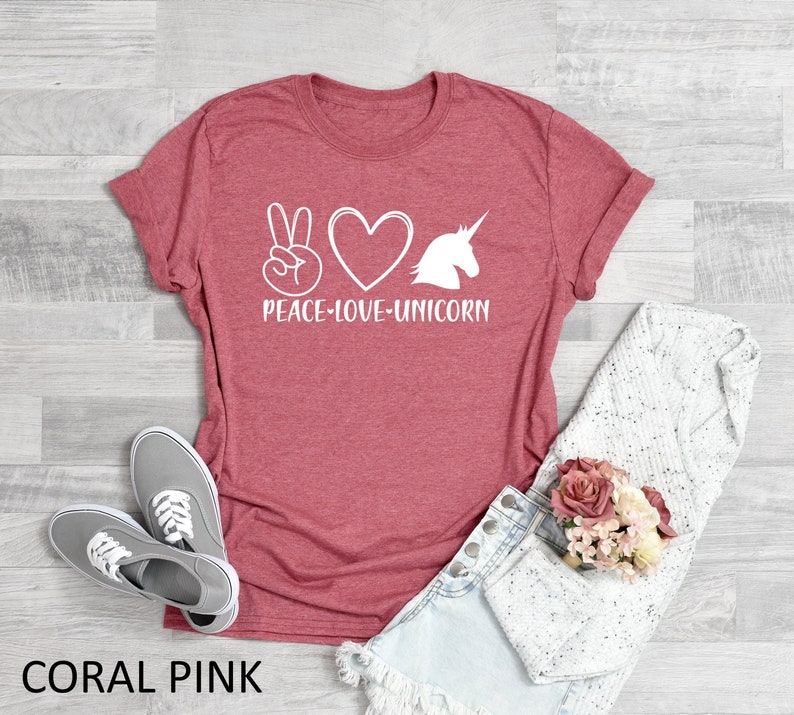 May include: Coral pink t-shirt with the words "Peace Love Unicorn" in white. The design includes a peace sign, a heart, and a unicorn silhouette. The shirt is paired with gray sneakers and denim shorts.