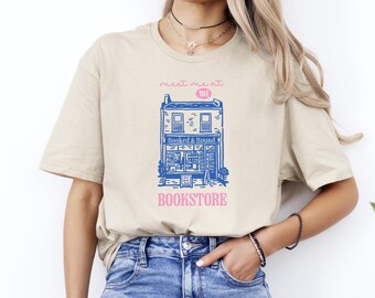 May include: Beige t-shirt featuring a blue and pink bookstore graphic. The graphic includes the text "meet me at the THE BOOKSTORE" and an illustration of a building with the words "Booked & Bound".