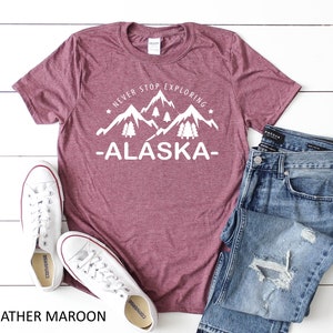 May include: Heather maroon t-shirt with white text that reads "Never Stop Exploring -Alaska-." The text is accompanied by a graphic of mountains and pine trees. The t-shirt is laid flat on a white wooden surface with a pair of white sneakers and blue jeans.