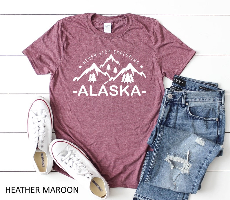 May include: Heather maroon t-shirt with white text that reads "Never Stop Exploring -Alaska-." The text is accompanied by a graphic of mountains and pine trees. The t-shirt is laid flat on a white wooden surface with a pair of white sneakers and blue jeans.