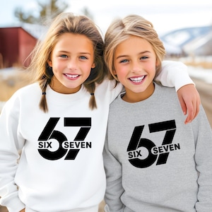 May include: Two young people wearing matching sweatshirts. One sweatshirt is white, the other is gray. Both sweatshirts have the number "67" in large black letters, with the words "SIX SEVEN" below.
