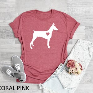 May include: Coral pink t-shirt with a white silhouette of a Doberman Pinscher with a heart in its chest. The shirt is laying flat on a woodgrain surface.