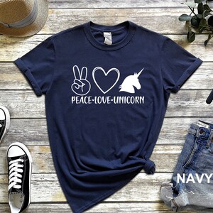 May include: Navy blue t-shirt with a white graphic design featuring a peace sign, a heart, and a unicorn silhouette, with the words "PEACE-LOVE-UNICORN" underneath. The shirt is folded and has rolled-up sleeves.