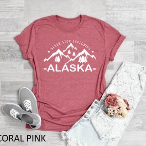 May include: Coral pink t-shirt with a white graphic of mountains and trees with the text "Never Stop Exploring -Alaska-"