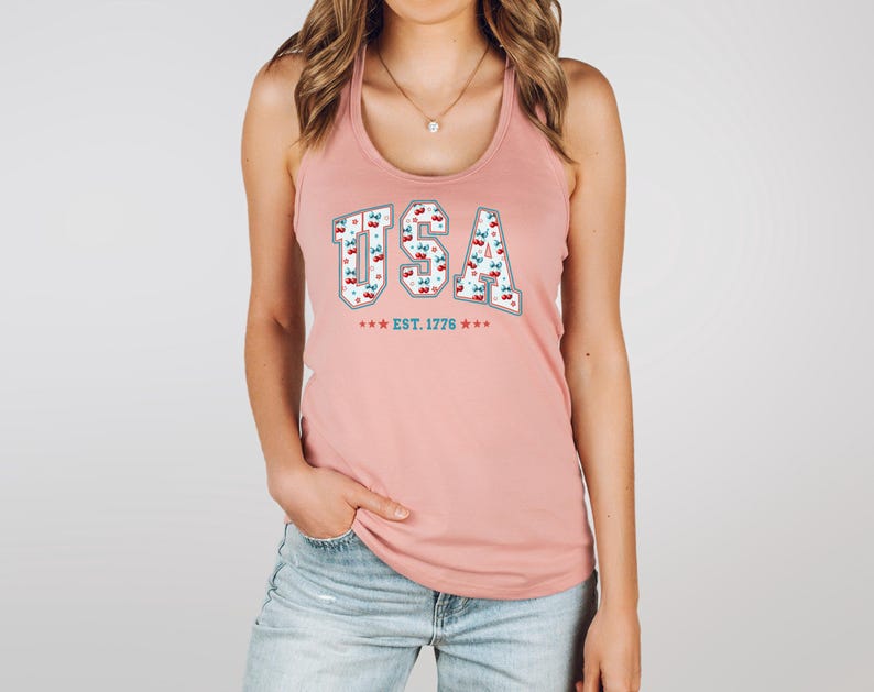 May include: A light pink tank top with the word "USA" in a white and gray font, filled with a cherry pattern. Below the text are three blue stars and the text "EST. 1776".
