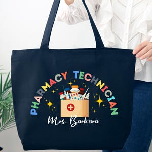 May include: Navy blue tote bag with the words "PHARMACY TECHNICIAN" in a rainbow arc above a box of medical supplies. The name "Mrs. Barbara" is printed below the box. The bag has two black handles.