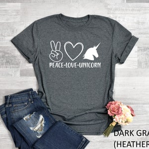 May include: Dark gray t-shirt with the words "PEACE-LOVE-UNICORN" in white. The design includes a peace sign, a heart, and a unicorn silhouette. The shirt is paired with blue jeans and a bouquet of pink flowers.