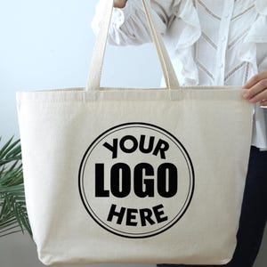 K&ouml;nnte beinhalten: Cremefarbene Tragetasche mit langen Henkeln. Auf der Vorderseite ist ein schwarzes kreisf&ouml;rmiges Logo mit dem Text "YOUR LOGO HERE" aufgedruckt. Die Tasche wird von einer Person gehalten.