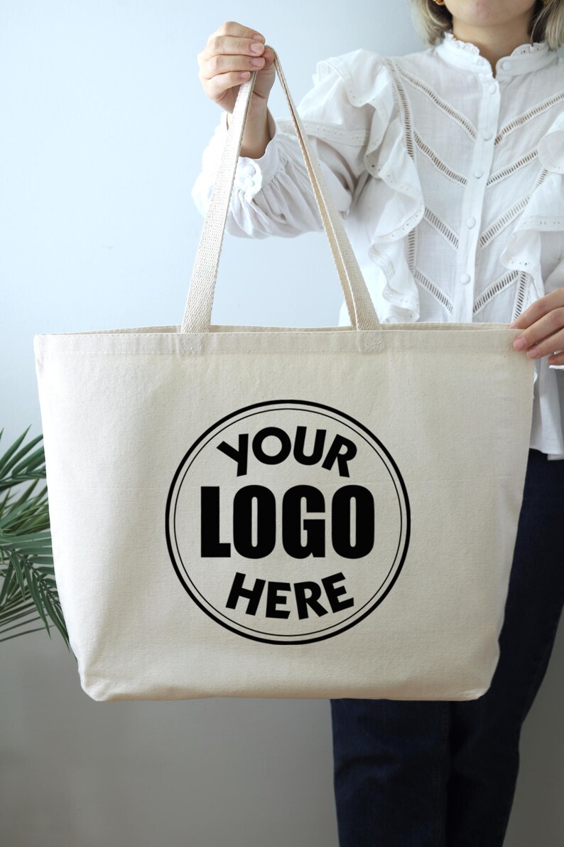 K&ouml;nnte beinhalten: Cremefarbene Tragetasche mit langen Henkeln. Auf der Vorderseite ist ein schwarzes kreisf&ouml;rmiges Logo mit dem Text "YOUR LOGO HERE" aufgedruckt. Die Tasche wird von einer Person gehalten.