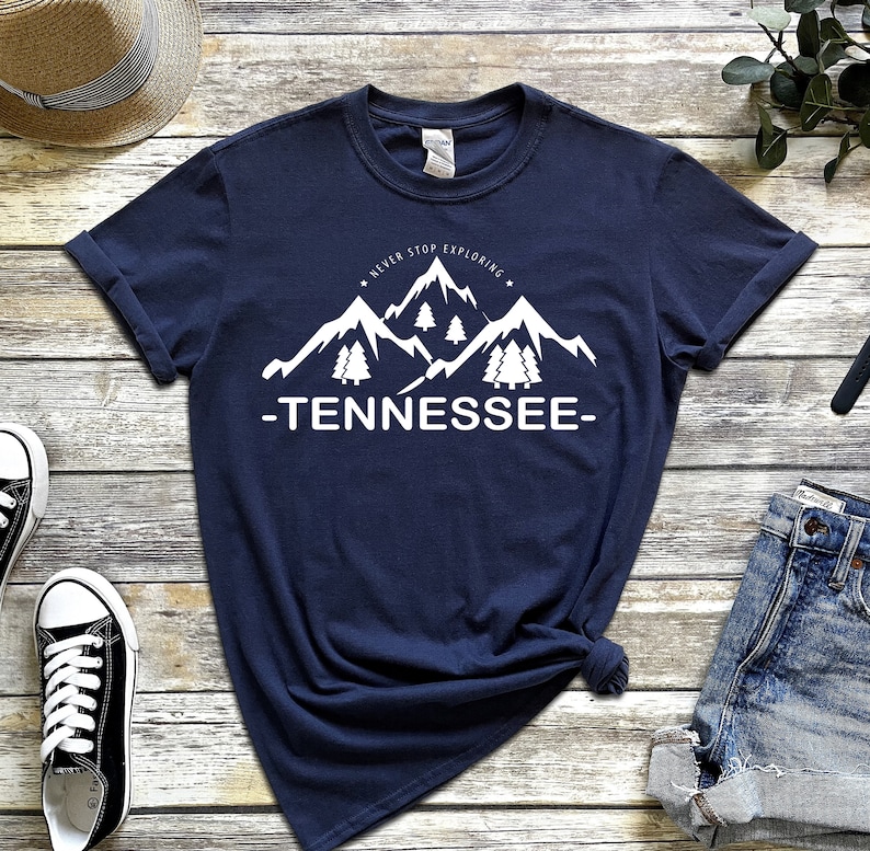 May include: A navy blue t-shirt with a white graphic of mountains and trees. The text "Never Stop Exploring" and "Tennessee" are printed on the shirt.