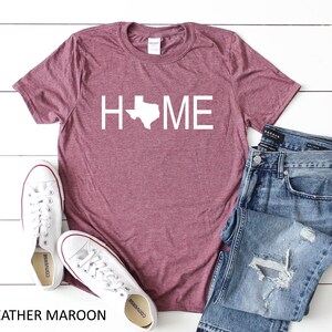 May include: Heather maroon t-shirt with a white outline of the state of Texas and the word "HOME" printed on the front. The shirt is paired with blue jeans and white sneakers.