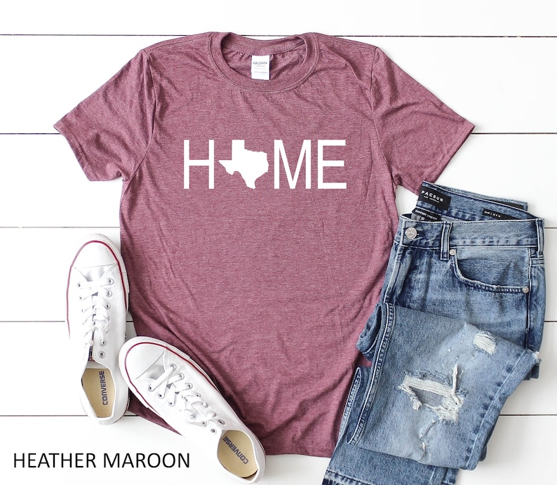 May include: Heather maroon t-shirt with a white outline of the state of Texas and the word "HOME" printed on the front. The shirt is paired with blue jeans and white sneakers.