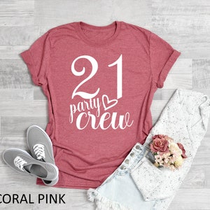 May include: Coral pink t-shirt with the text "21 party crew" in white script. The shirt is paired with denim shorts, gray sneakers, and a white sweater. A small bouquet of pink and cream flowers is also present.