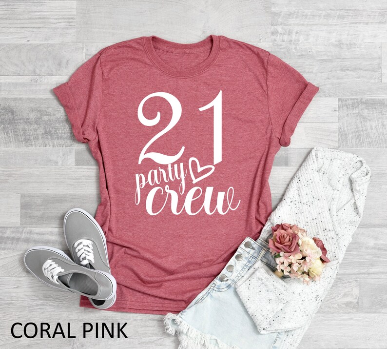 May include: Coral pink t-shirt with the text "21 party crew" in white script. The shirt is paired with denim shorts, gray sneakers, and a white sweater. A small bouquet of pink and cream flowers is also present.