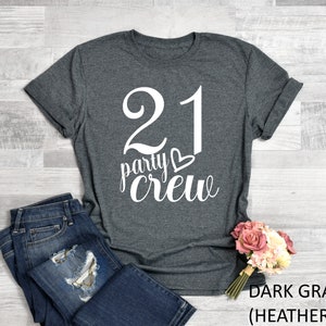 May include: Dark gray heather t-shirt with the text "21 party crew" in white script. The number 21 is large and bold. A pair of blue jeans and a bouquet of pink and white flowers are next to the shirt.