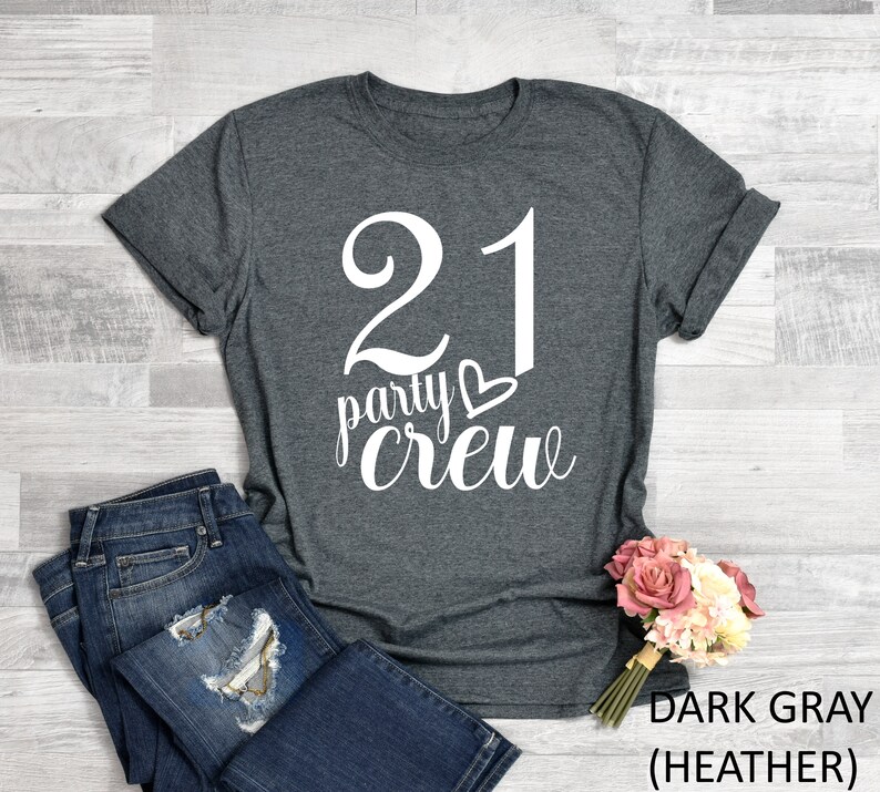May include: Dark gray heather t-shirt with the text "21 party crew" in white script. The number 21 is large and bold. A pair of blue jeans and a bouquet of pink and white flowers are next to the shirt.