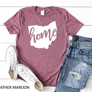 K&ouml;nnte beinhalten: Heather-marronfarbenes T-Shirt mit einer wei&szlig;en Umrandung des Bundesstaates Ohio und dem Wort "home" in wei&szlig;er, kursiv geschriebener Schrift. Das Shirt liegt auf einer wei&szlig;en Holzoberfl&auml;che mit einem Paar wei&szlig;en Turnschuhen und einer blauen Jeans.