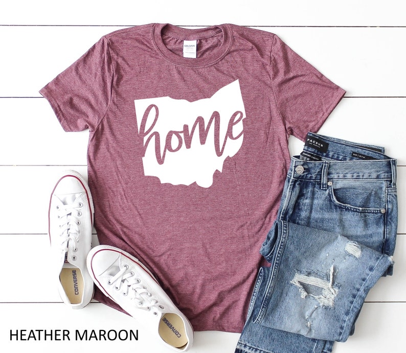 K&ouml;nnte beinhalten: Heather-marronfarbenes T-Shirt mit einer wei&szlig;en Umrandung des Bundesstaates Ohio und dem Wort "home" in wei&szlig;er, kursiv geschriebener Schrift. Das Shirt liegt auf einer wei&szlig;en Holzoberfl&auml;che mit einem Paar wei&szlig;en Turnschuhen und einer blauen Jeans.