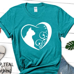 May include: Teal blue t-shirt with a white silhouette of a cat inside a heart with floral designs. The text "DEEP TEAL (Heather)" is printed below the image.