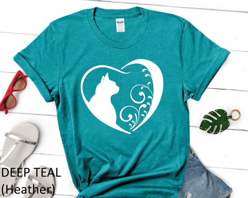 May include: Teal blue t-shirt with a white silhouette of a cat inside a heart with floral designs. The text "DEEP TEAL (Heather)" is printed below the image.