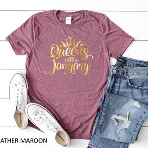 May include: Heather maroon t-shirt with gold glitter lettering that says "Queens are born in January". The shirt is shown on a white surface with a pair of white sneakers and blue jeans.