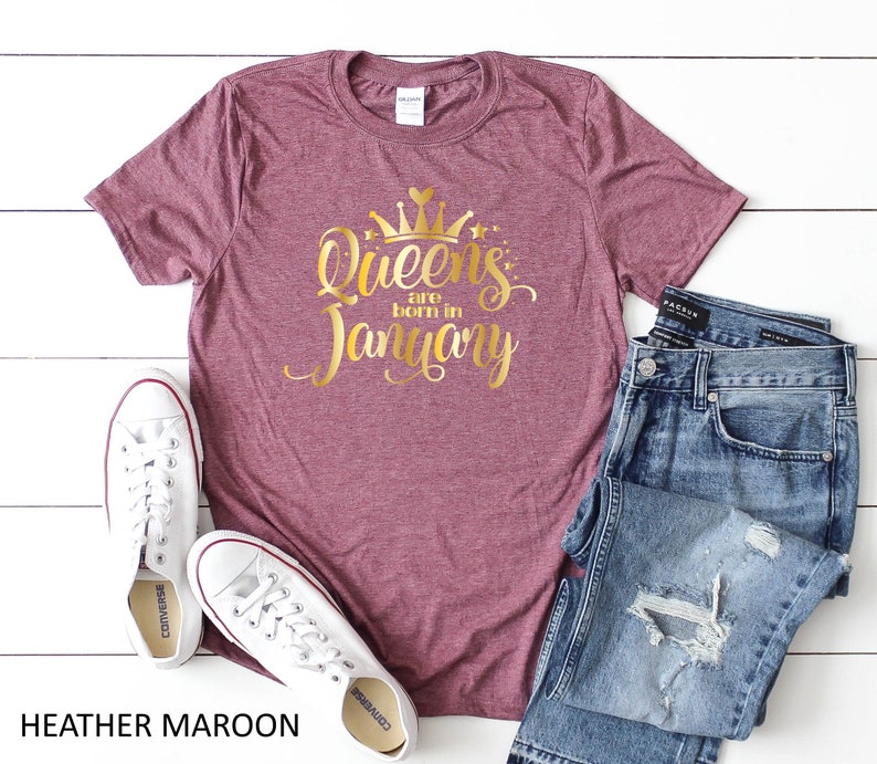 May include: Heather maroon t-shirt with gold glitter lettering that says "Queens are born in January". The shirt is shown on a white surface with a pair of white sneakers and blue jeans.