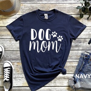 May include: Navy blue t-shirt with the words "DOG mom" in white script, accompanied by two white paw prints. The shirt is folded at the bottom and is paired with denim shorts. The word "NAVY" is visible on the shorts.