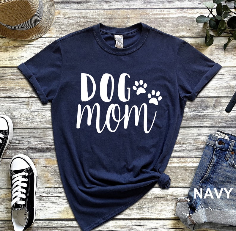 May include: Navy blue t-shirt with the words "DOG mom" in white script, accompanied by two white paw prints. The shirt is folded at the bottom and is paired with denim shorts. The word "NAVY" is visible on the shorts.