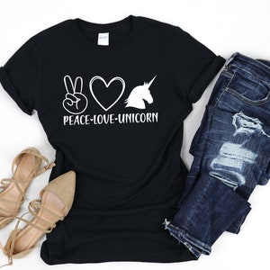 May include: Black t-shirt with the words "PEACE-LOVE-UNICORN" in white. The design includes a peace sign, a heart, and a unicorn silhouette. The shirt is paired with blue jeans and beige sandals.