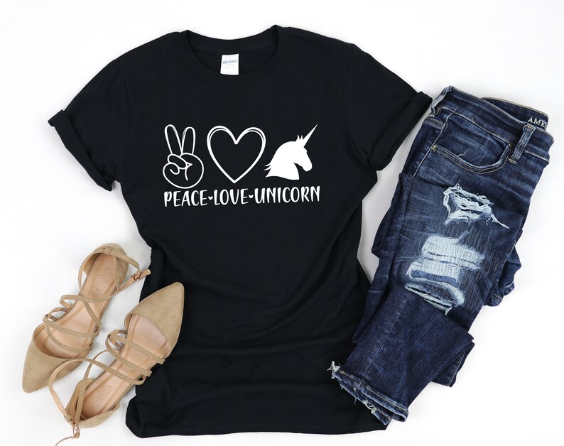 May include: Black t-shirt with the words "PEACE-LOVE-UNICORN" in white. The design includes a peace sign, a heart, and a unicorn silhouette. The shirt is paired with blue jeans and beige sandals.